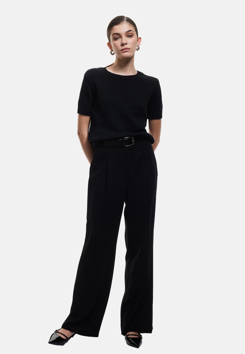 Sort, kortærmet striktrøje sat sammen med højtaljede, vide sorte bukser og spidse sorte flade sko. Bælte tilføjer struktur.