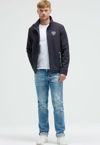 Chaqueta softshell de color azul marino oscuro con un cuello alto, bolsillos con cremallera y un parche con el logo. Llevada sobre una camiseta blanca y combinada con jeans de color azul claro.