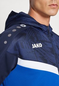 Navy blue athletic hoodie with a textured pattern, white accents, and a logo. Features zipper closure and round decorative dots on the shoulder.