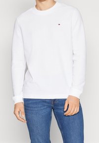 Vit långärmad t-shirt med rund halsringning och ribbad textur samt en liten logotyp på bröstet, tillsammans med blå denimjeans.