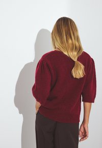 Burgundi spalvos megztinis su pūstomis trumpomis rankovėmis ir laisvu siluetu. Tekstūruota medžiaga su raukšlėmis apačioje ir ant rankovių. Nešiojamas su tamsiomis kelnėmis.