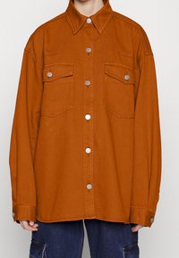 Une chemise en jean orange surdimensionnée, avec un col pointu, deux poches poitrine, des accessoires en argent et des fermetures à boutons sur le devant.