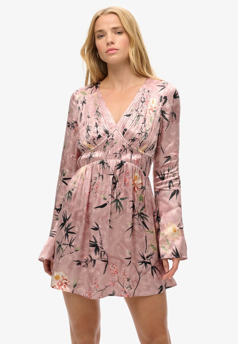 Vestido corto rosa hecho de una tela sedosa, con escote en V, mangas largas y un patrón floral en tonos negros y amarillos.