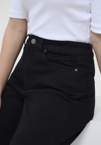 Black high-waisted jeans with front pockets, a fitted design, and metal accents. The fabric shows a smooth, slightly textured finish.