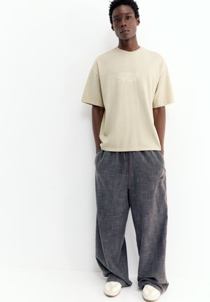 Giovane persona che indossa una t-shirt beige con scritta e pantaloni grigi larghi, in piedi davanti a uno sfondo bianco uniforme con le mani nelle tasche.