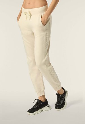 Beige Sweatpants mit seitlichen Nähten und strukturierten Akzenten, elastischem Bund mit Kordelzug und Seitentaschen, kombiniert mit schwarzen Sneakers.
