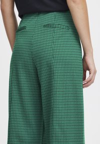 Pantaloni a quadri verdi con un tessuto testurizzato, dotati di due tasche posteriori e un design a vita alta. Il motivo è composto da quadrati neri e verdi.