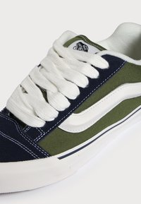 Baskets basses Vans en toile bleu marine et vert olive, bande latérale blanche, lacets blancs épais et semelle en caoutchouc blanche.