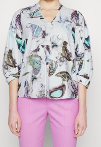 Blusa azzurro chiaro con stampe di farfalle e fiori. Ha un colletto a V, chiusura con bottoni e maniche a tre quarti. Abbinata a pantaloni rosa.