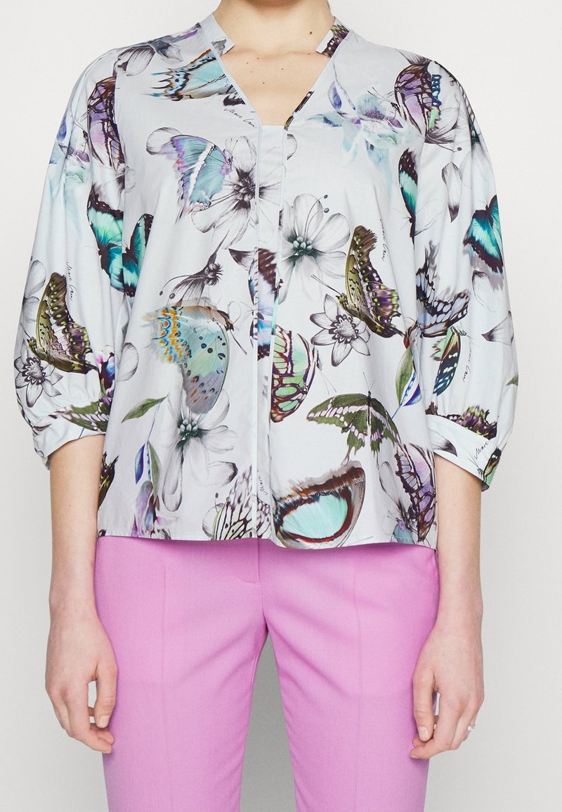Blusa azzurro chiaro con stampe di farfalle e fiori. Ha un colletto a V, chiusura con bottoni e maniche a tre quarti. Abbinata a pantaloni rosa.