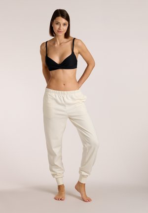 Woman standing barefoot wearing a black bra and off-white jogger pants against a plain beige background.