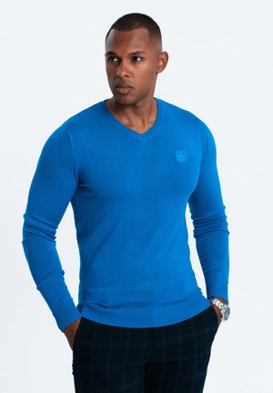 Pull bleu à manches longues en V, fabriqué dans un tissu doux, avec des poignets et un ourlet côtelés. Porté avec un pantalon à motif écossais.