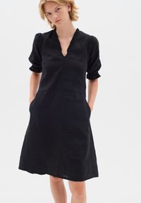 Schwarzes Leinenkleid mit kurzen Puffärmeln, V-Ausschnitt, Rüschendetails am Ausschnitt und seitlichen Taschen. Eine entspannte A-Linien-Silhouette.