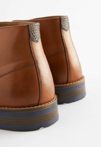 Botas de cuero marrón hasta el tobillo con acentos grises texturizados y una suela de goma azul, presentando un acabado suave y detalles de costura en contraste.