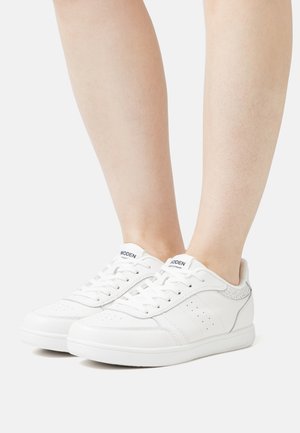 Sneakers bianchi con dettagli perforati e accenti grigi texturizzati. Presentano lacci piatti e una suola imbottita, esposti su una gamba nuda.