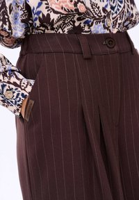 Bruine pinstripe broeken met een getailleerde pasvorm, voorzien van zijzakken en een knoopsluiting. Aangevuld met een stoffen label en een gestructureerd materiaal.