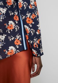 Blusa floreale in tessuto blu navy con fiori arancioni e bianchi. Bordo con spacchi laterali in azzurro chiaro, polsini con bottoni e texture morbida.