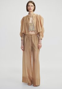 Beige doorschijnende jas met opgezette mouwen, over een met pailletten bezette crop top, gecombineerd met losse, elastische brede pijpenbroek. Gladde textuur overal.