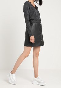 Jupe en similicuir noir à taille haute, avec des boutons sur le devant et une texture lisse, associée à un haut à manches longues foncé et des baskets blanches.