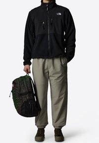 Czarna polarowa kurtka z kieszeniami na suwak; beżowe luźne spodnie; zielono-czarny plecak w wzory; brązowe buty do wędrówek.