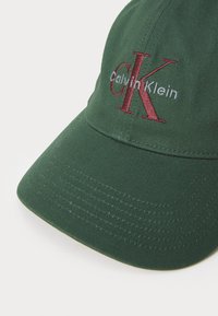 Groene katoenen pet met een gebogen klep. Bevat geborduurd "Calvin Klein" logo in rood en wit, met textuurstof.