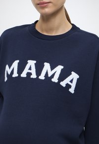 Μπλε σκούρο φούτερ με λευκά, αφράτα γράμματα "MAMA". Διαθέτει στρογγυλή λαιμόκοψη και απαλή υφή υφάσματος, κομμένο λίγο πάνω από τη μέση.