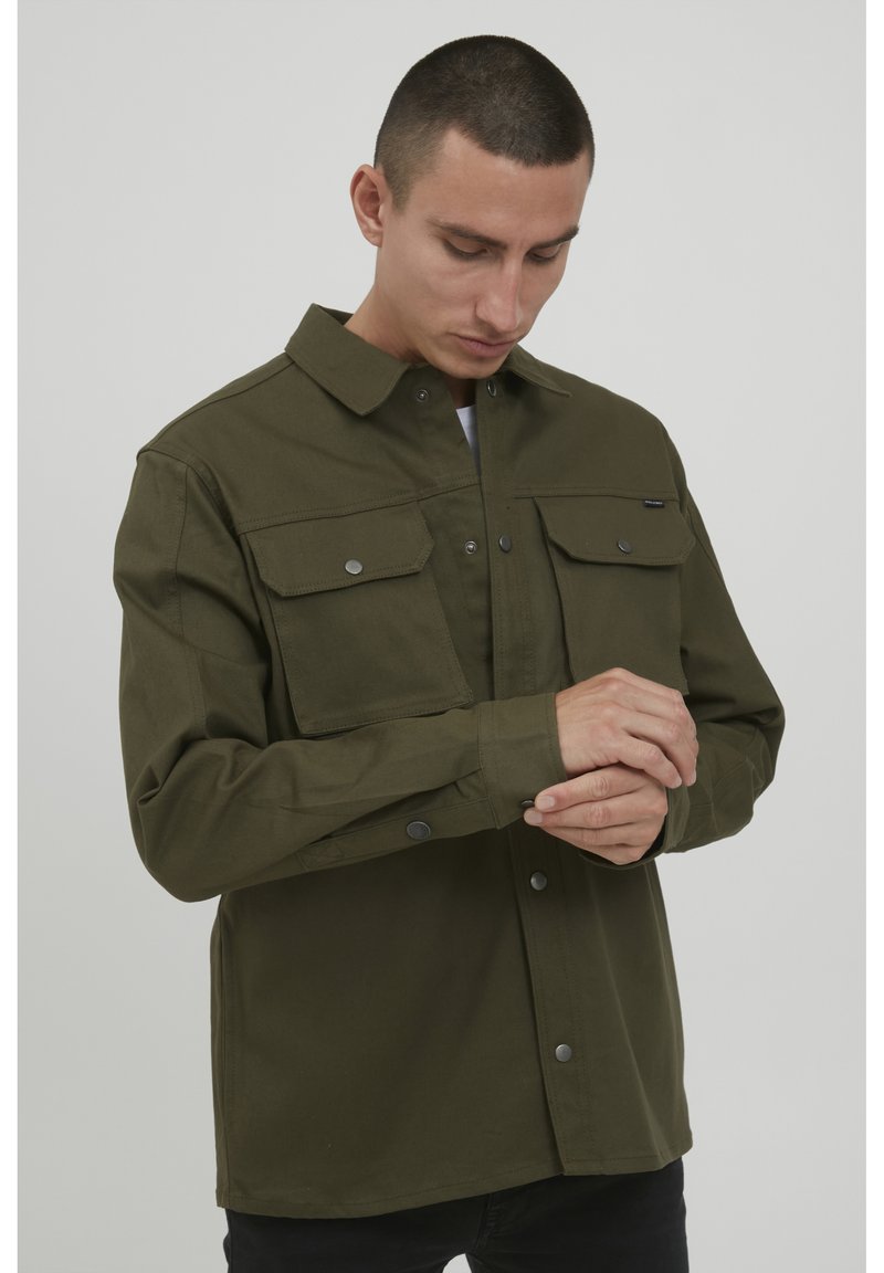 Camisa verde oliva de botón, hecha de una tela resistente, con dos bolsillos en el pecho y botones a presión, que presenta un cuello clásico y mangas largas.
