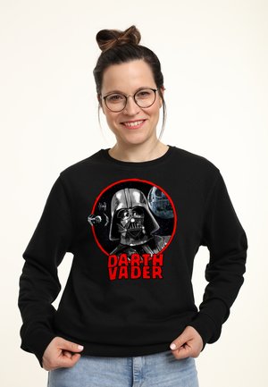 Smilende kvinde med briller iført sort sweatshirt med Darth Vader-hjelm, Dødsstjernen og TIE-fighter i en rød cirkel med teksten "DARTH VADER".