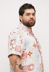 Chemise à manches courtes en tissu léger avec un design en patchwork coloré comprenant des motifs bleus, rouges, oranges et crème. Col à boutons.