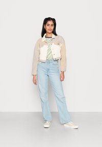 Casaco de ganga beige e branco cortado, por cima de uma camisa riscada verde e branca, combinado com jeans largos azul claro e ténis claros.