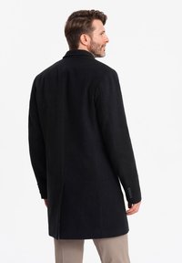 Abrigo de lana negro con un corte entallado, mangas largas, collar con muescas y acentos de puños abotonados. Textura suave; diseño hasta la rodilla.