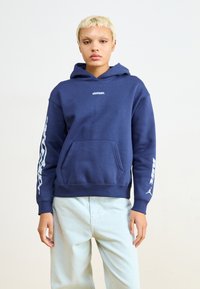 Sweatshirt azul-marinho com bolso frontal, punhos canelados e texto gráfico nas mangas. Material de fleece macio, corte descontraído e capuz.
