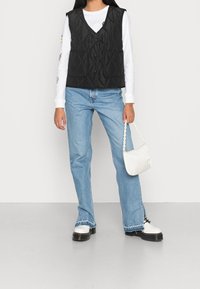 Svart quiltad väst med två framfickor, lager över en vit långärmad skjorta. Ljusblå raka jeans och en vit handväska.