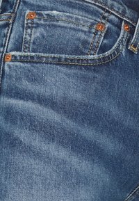 Denim fabric in dark blue with a slight wash effect. Features copper rivets and stitching on pockets, highlighting a classic jeans design.