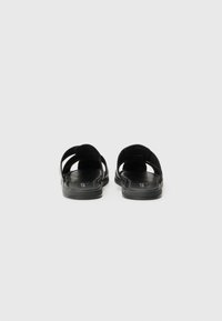 Black sandals with a flat sole, featuring two wide straps that cross over the top and open sides for ventilation. Smooth texture throughout.