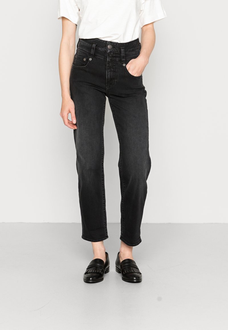 Femme portant un jean noir taille haute à jambes droites, un haut blanc à manches courtes et des mocassins en cuir noirs, debout avec une main dans la poche.