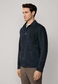 Marineblauwe suède jas met een kraag, knoopsluiting en twee voorkruiken; gestructureerde afwerking geaccentueerd door zichtbare stiksels.