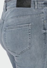 Jeans de mezclilla azul claro, con corte slim, bolsillo trasero y un parche de cuero negro texturizado en la cintura.
