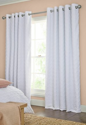 Tende bianche strutturate pendono da un’asta di metallo in una camera da letto beige con una finestra, un letto con un cuscino e un tappeto intrecciato su pavimento in legno.