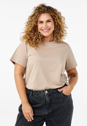 Helles beige T-Shirt mit kurzen Ärmeln und rundem Ausschnitt, kombiniert mit hochgeschnittenen, dunklen Jeans. Das Model lächelt beim Posieren.