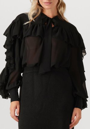 Vrouw in een doorzichtige zwarte blouse met gelaagde ruffles op de mouwen en schouders, met een strik om de nek, gecombineerd met een zwarte hoge taille rok.
