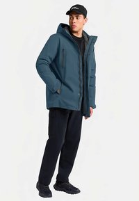 Waterdichte jas in turquoise met capuchon, voorste drukknoopsluiting en twee met rits afsluitbare zakken. Draagt zwarte broek en sneakers. Eenvoudig, functioneel ontwerp.