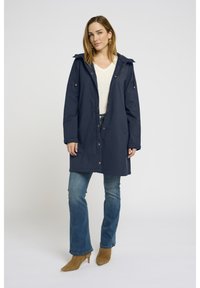 Manteau long marine avec capuche à cordon, fermeture à boutons-pression et poches latérales, associé à un jean clair et des bottines beiges.