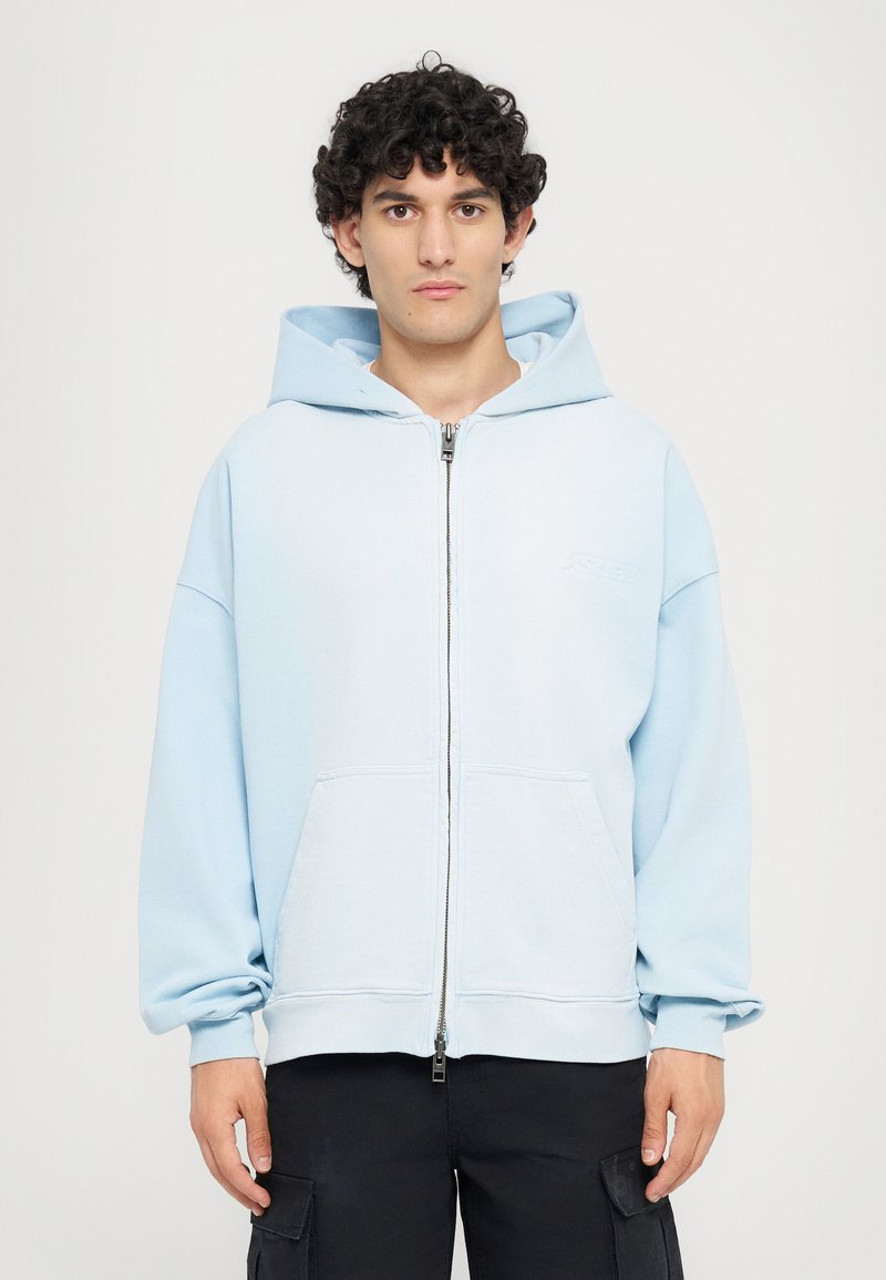 Gaiši zilā zip-up džemperis ar lielu priekšējo kabatu, ribotām piedurknēm un oderētu kapuci. Uz krūtīm ir neliels logotips. Mīksta auduma tekstūra.