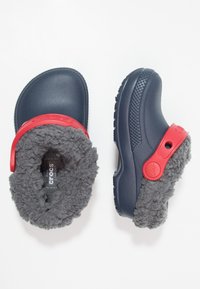 Granatowe chodaki z miękkim szarym futrzanym wykończeniem i czerwonym paskiem. Posiadają teksturowaną podeszwę i zaokrąglony kształt noska.
