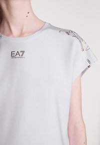 Jasnoszary t-shirt z zaokrąglonym dekoltem, krótkimi rękawami z rolowanym wykończeniem oraz złotymi akcentami na ramionach. Zawiera logo EA7 w kolorze brązowym.