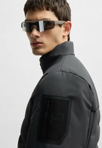 Svart pufferjacka med hög krage, med en ficka med blixtlås på vänster arm. Modell som bär grå sportsolglasögon med spegelglas.