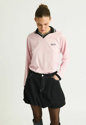 Kvinde med håret samlet bagpå iført en lyserød bluse med lange ærmer og sort krave, sort ballon-nederdel, sorte strømpebukser og sorte støvler.