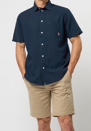 Homme portant une chemise bleu marine à manches courtes avec poche avant et un short beige à la longueur genou, mains dans les poches, sur fond uni.