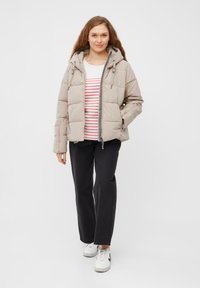 Beige pufferjack met een capuchon, rits aan de voorkant en elastische details, gedragen over een wit-rood gestreept shirt en zwarte broek. Witte sneakers zijn zichtbaar.